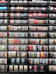 A large bookshelf filled with a variety of books stacked neatly in rows. The books have colorful spines with a mix of titles and authors visible. The shelf is black, creating a contrast with the diverse colors of the books.