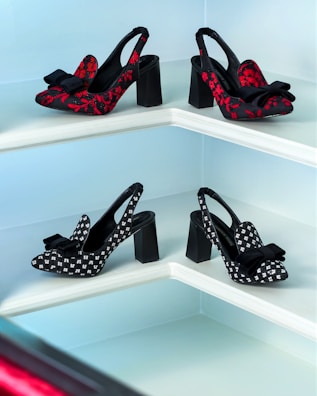 A display of four pairs of high-heeled shoes arranged on a tiered white shelf. The shoes on the top row are decorated with red floral patterns and black bows, while the pairs on the bottom row feature black and white patterns with black bows. The shelves are set against a light blue background.