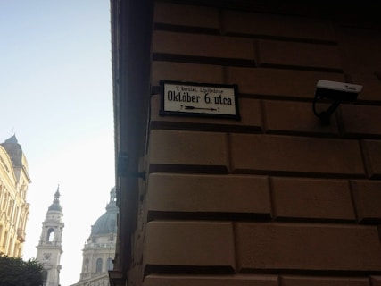 A street sign on a building reads 'Október 6. utca', accompanied by an arrow. A surveillance camera is mounted on the wall next to the sign. Architectural elements of an ornate building and domed tower are visible, silhouetted against a bright sky.