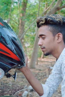 A person with styled hair is closely observing a motorcycle helmet. The helmet is black with red accents, and the background is a lush forest area.