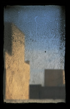 A frosted glass window with abstract shapes and colors. The blurred outlines of structures cast shadows, creating a soft blend of warm and cool hues.