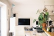 A neatly organized home office space features a large computer monitor on a wooden desk. There is a white ergonomic chair in front of the desk, a tall potted plant to the right, and kitchen utensils hanging above a sink area. An anglepoise lamp is positioned next to some filing drawers.