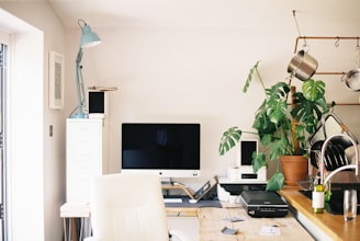 A neatly organized home office space features a large computer monitor on a wooden desk. There is a white ergonomic chair in front of the desk, a tall potted plant to the right, and kitchen utensils hanging above a sink area. An anglepoise lamp is positioned next to some filing drawers.