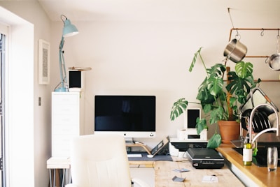 A neatly organized home office space features a large computer monitor on a wooden desk. There is a white ergonomic chair in front of the desk, a tall potted plant to the right, and kitchen utensils hanging above a sink area. An anglepoise lamp is positioned next to some filing drawers.
