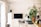 A neatly organized home office space features a large computer monitor on a wooden desk. There is a white ergonomic chair in front of the desk, a tall potted plant to the right, and kitchen utensils hanging above a sink area. An anglepoise lamp is positioned next to some filing drawers.