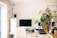 A neatly organized home office space features a large computer monitor on a wooden desk. There is a white ergonomic chair in front of the desk, a tall potted plant to the right, and kitchen utensils hanging above a sink area. An anglepoise lamp is positioned next to some filing drawers.