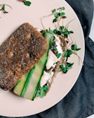 A sandwich made with dark whole grain bread, filled with fresh green sprouts, cucumber slices, and a creamy spread on a light-colored plate, set against a dark fabric backdrop.