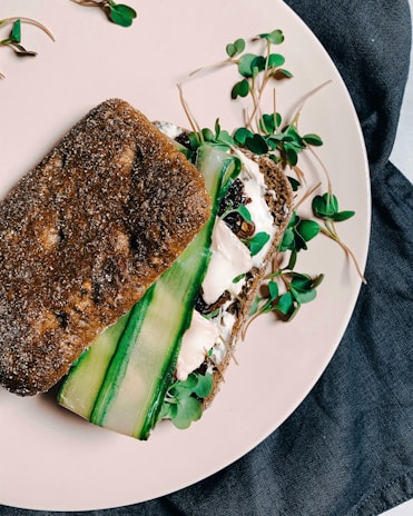 A sandwich made with dark whole grain bread, filled with fresh green sprouts, cucumber slices, and a creamy spread on a light-colored plate, set against a dark fabric backdrop.