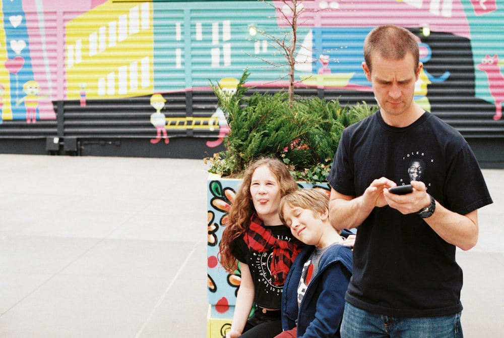man standing and using phone near two children sitting
