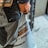 A person is wearing orange safety gloves and holding a white construction bag. The bag has text written on it, partially visible, and the person appears to be dressed in work overalls with a dark color on top and grayish-blue on the bottom.