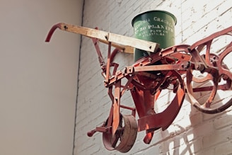 A vintage agricultural machine is mounted on a white brick wall. The machinery has rusted red metal parts with gears, wheels, and wooden handles. A green metal container with writing is attached to the top.