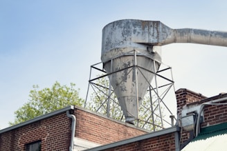 A large, weathered industrial funnel or silo is mounted on top of a brick building. The funnel has a rusty, aged appearance, with exposed metal and peeling paint. It's supported by a metal framework. To the right, a large horizontal pipe extends from the top of the funnel. In the background, foliage from a tree can be seen against a clear blue sky.