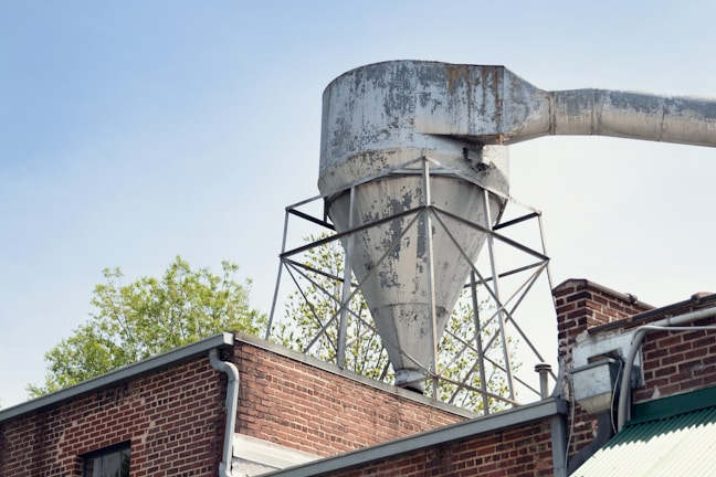 A large, weathered industrial funnel or silo is mounted on top of a brick building. The funnel has a rusty, aged appearance, with exposed metal and peeling paint. It's supported by a metal framework. To the right, a large horizontal pipe extends from the top of the funnel. In the background, foliage from a tree can be seen against a clear blue sky.