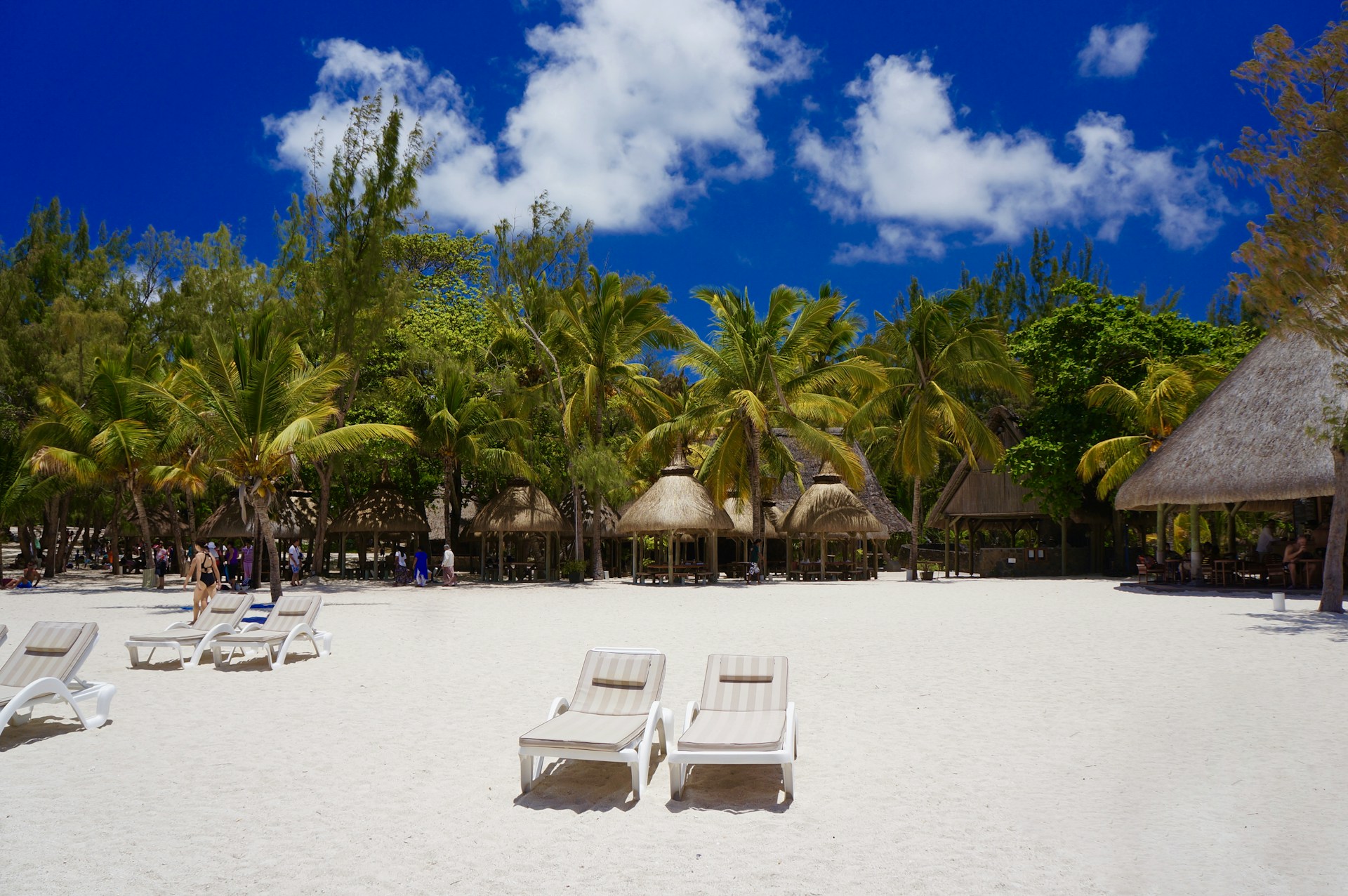 A tropical beach with white sand, lined with palm trees and thatched roof huts. The clear blue sky is dotted with a few clouds. Several lounge chairs are arranged on the sand, some of which are occupied by people.