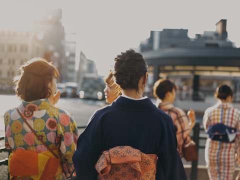 A group of people are wearing traditional kimonos, with their backs facing the camera. The scene is set outdoors on a sunny day, with soft lighting and blurred urban buildings in the background. Each kimono features different patterns and colors, adding vibrancy to the image.