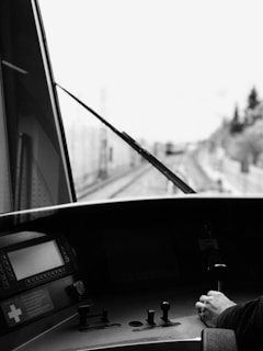 A black and white photograph taken from inside a train cab. The focus is on the control panel with various knobs and a display screen, alongside a person's hand gripping a control lever. The blurred view through the windshield shows railway tracks and distant trees.
