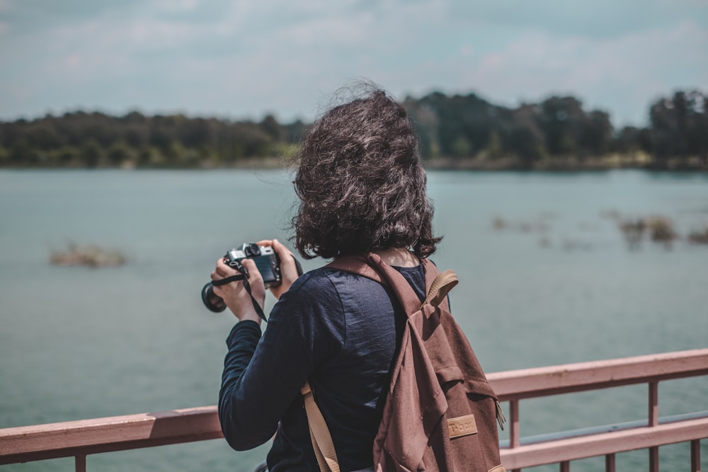 woman wearing black shirt and brown backpack holding camera standing near railings near body of water