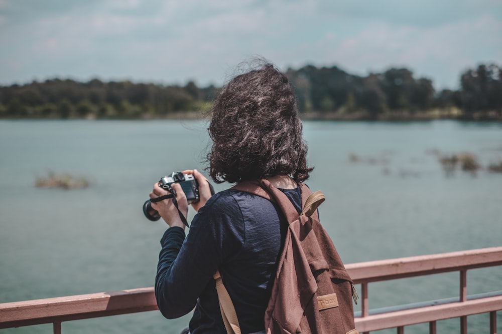 Frau mit schwarzem Hemd und braunem Rucksack mit Kamera steht in der Nähe eines Geländers in der Nähe eines Gewässers