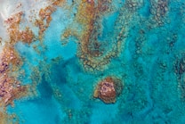 Aerial view of a vibrant and clear turquoise ocean, scattered with rocky formations and coral reefs. The water's shades range from deep blue to light aqua, interspersed with brown and orange hues from the rocks.