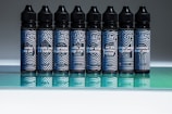 Seven bottles with intricate geometric patterns stand in a row, each with distinct phrases such as 'loves you all' and 'breaks your old habits' printed on the labels. The design features primarily black and white stripes with blue accents, set against a reflective green surface.
