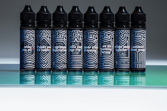 Seven bottles with intricate geometric patterns stand in a row, each with distinct phrases such as 'loves you all' and 'breaks your old habits' printed on the labels. The design features primarily black and white stripes with blue accents, set against a reflective green surface.