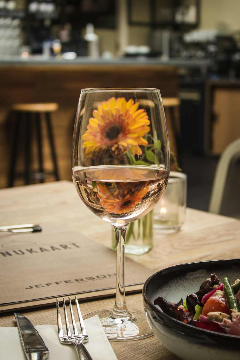 Weißwein auf Glas in der Nähe von Gabel und Messer aus grauem Edelstahl neben orangefarbener Gerbera-Gänseblümchenblüte auf dem Tisch
