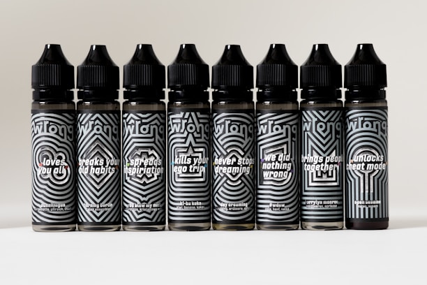 Seven black bottles with black and white geometric designs and text are arranged in a row. Each bottle features a unique phrase such as 'loves you all,' 'breaks your old habits,' and 'spreads inspiration.'