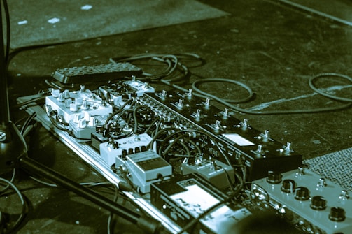 A collection of guitar effect pedals arranged on a pedalboard with various cables and connections. The setup includes multiple pedals of different sizes and types, organized neatly. The background shows a dark floor with scattered small particles and a textured mat.