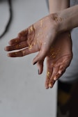 Two hands are open and covered with scattered gold glitter. The skin tone is light, and the nails are painted in dark colors. The background is blurred, adding focus to the hands.