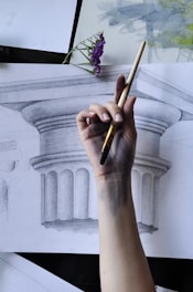 A hand holding a paintbrush is positioned over a detailed pencil sketch of a classic architectural column. The fingers are stained with graphite, suggesting recent or ongoing art work. Above the sketch, a small purple flower adds a touch of color and natural contrast. The scene combines elements of creative expression and classical art.