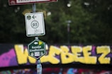 A close-up of multiple road signs attached to a post in front of a graffiti-covered wall. The upper sign indicates a bike path with a number 95 and an arrow pointing left. Below it, a green sign says 'Rondje Eindhoven.' The background features bold yellow graffiti against a dark surface with a mix of other colors.