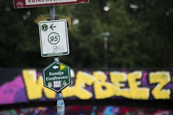 A close-up of multiple road signs attached to a post in front of a graffiti-covered wall. The upper sign indicates a bike path with a number 95 and an arrow pointing left. Below it, a green sign says 'Rondje Eindhoven.' The background features bold yellow graffiti against a dark surface with a mix of other colors.