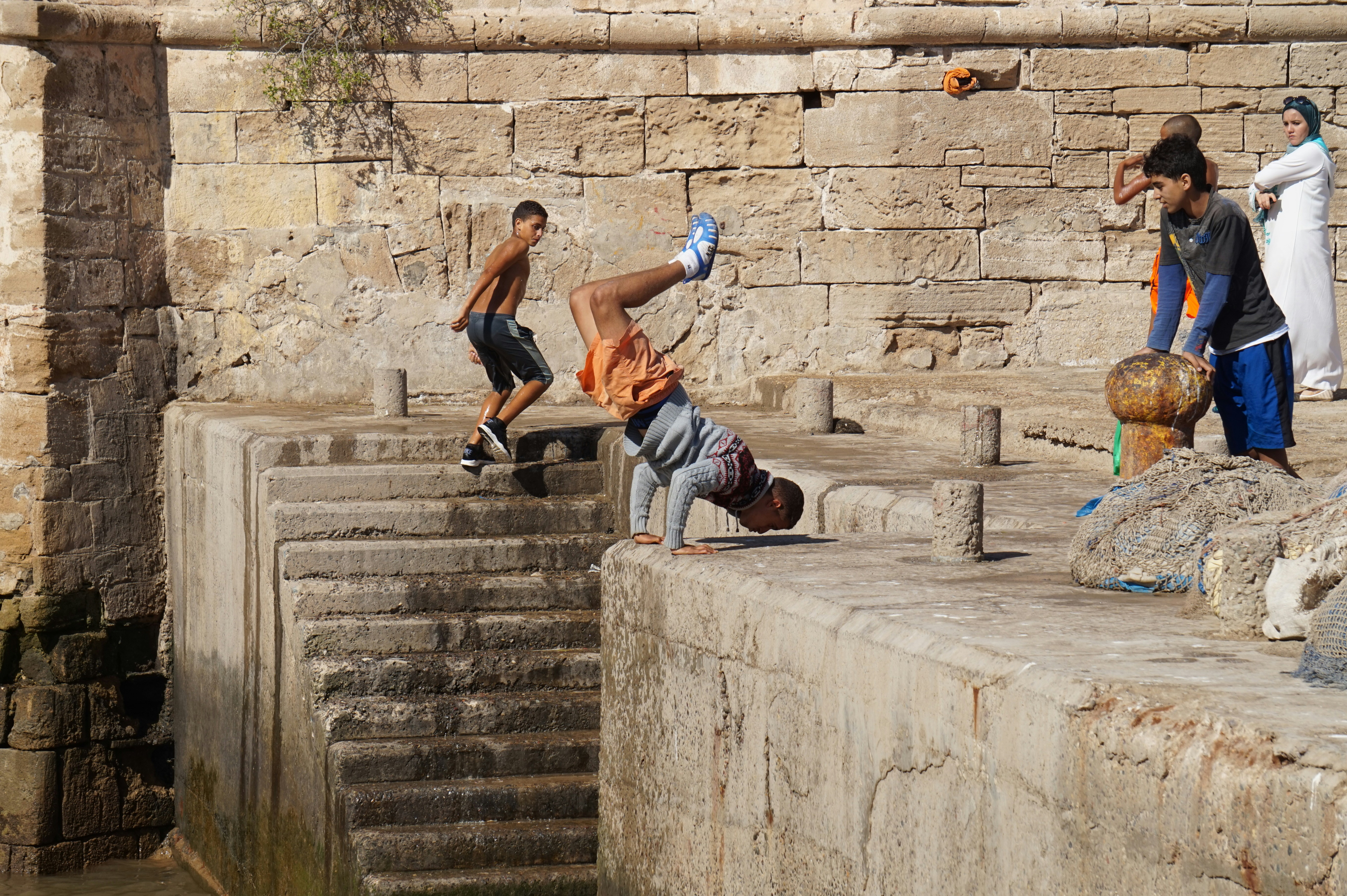 Photograph of a youth mid-flip from a stone ledge, as onlookers watch along the water’s edge.