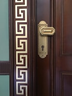 A close-up of a wooden door with an ornate brass door handle and keyhole. The door features intricate geometric designs in gold on the left side, giving it a classical and luxurious appearance.
