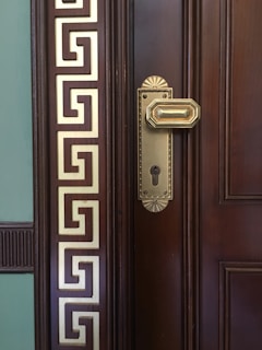 A close-up of a wooden door with an ornate brass door handle and keyhole. The door features intricate geometric designs in gold on the left side, giving it a classical and luxurious appearance.