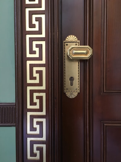 A close-up of a wooden door with an ornate brass door handle and keyhole. The door features intricate geometric designs in gold on the left side, giving it a classical and luxurious appearance.