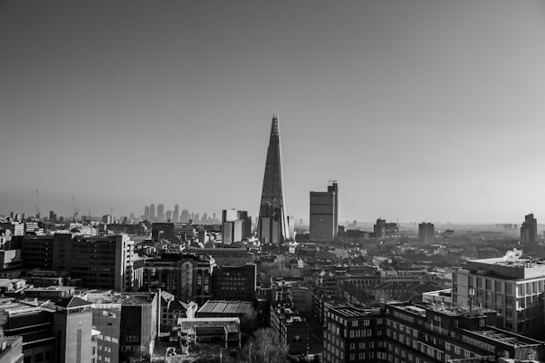 A black and white cityscape featuring numerous buildings with a prominent tall, modern skyscraper at the center, surrounded by smaller structures. The sky is clear with a gentle gradient from darker at the top to lighter near the horizon, where more distant buildings are visible, adding depth to the scene.