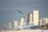 A bird is flying towards the camera, with a cityscape featuring tall buildings in the background. The focus is on the bird, giving it a sharp appearance against the blurred urban backdrop.