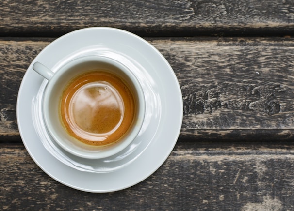 A white cup filled with espresso sits on a matching saucer. The cup rests on a wooden surface with a rustic texture, showcasing the rich, dark color of the coffee against the white ceramic.