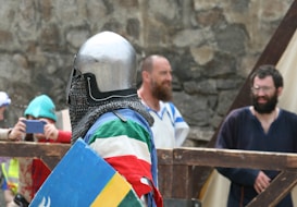 A person in medieval armor, including a helmet and chainmail, is holding a colorful shield with red, green, white, and blue sections. In the background, several people are present, some wearing costumes and others capturing the scene with a smartphone. The setting appears to be outdoors, possibly during a medieval reenactment event, with stone walls in the background.