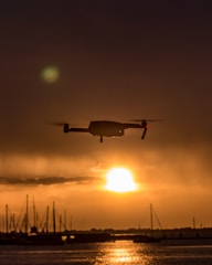 A drone is silhouetted against a vibrant orange sunset over a body of water. Sailboats and a horizon line can be seen in the background, creating a peaceful and scenic maritime landscape.