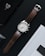 A brown leather-strap analog wristwatch with a white face is positioned on a dark surface alongside a smartphone and a book. The smartphone screen displays the time as 6:36 on Thursday, April 25. The book cover is predominantly white and partially visible.