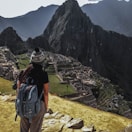 A person wearing a backpack and a knitted hat stands on a rocky foreground, overlooking the ancient ruins of Machu Picchu set against a backdrop of towering mountains. The scene conveys a sense of vastness and historical mystery.
