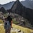A person wearing a backpack and a knitted hat stands on a rocky foreground, overlooking the ancient ruins of Machu Picchu set against a backdrop of towering mountains. The scene conveys a sense of vastness and historical mystery.