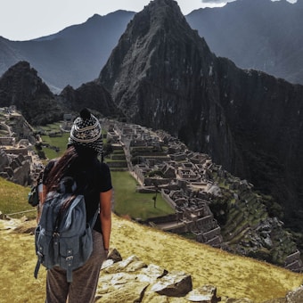A person wearing a backpack and a knitted hat stands on a rocky foreground, overlooking the ancient ruins of Machu Picchu set against a backdrop of towering mountains. The scene conveys a sense of vastness and historical mystery.