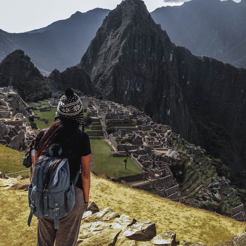 A person wearing a backpack and a knitted hat stands on a rocky foreground, overlooking the ancient ruins of Machu Picchu set against a backdrop of towering mountains. The scene conveys a sense of vastness and historical mystery.