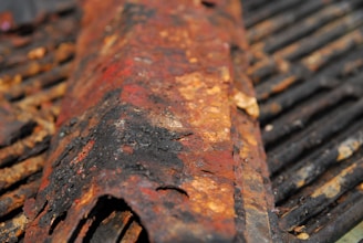 Close-up of a heavily rusted and corroded metal surface, showing flaking and uneven textures with various shades of red, brown, and orange. The background features metal bars that are also affected by rust.