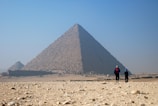 Two people walk towards the massive pyramids in a barren desert landscape under a clear blue sky. The pyramid’s ancient stone blocks are visible, casting shadows that accentuate its monumental structure.