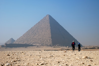 Two people walk towards the massive pyramids in a barren desert landscape under a clear blue sky. The pyramid’s ancient stone blocks are visible, casting shadows that accentuate its monumental structure.