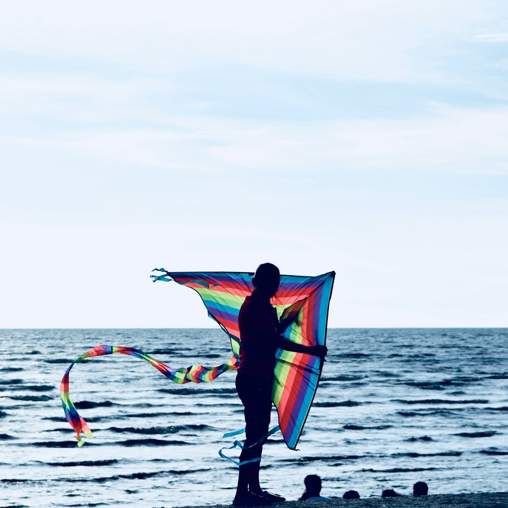 silhouette of man holding multicolored kite beside body of water during daytime